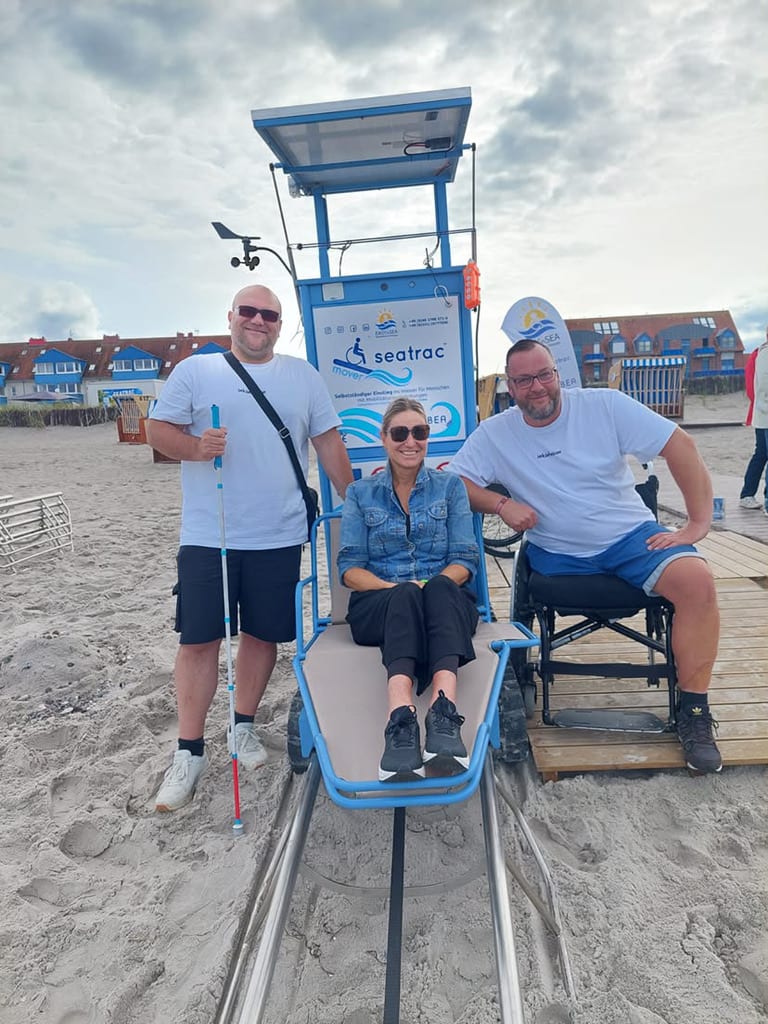 Kirsten Bruhn sitzt mit Sonnenbrille auf dem Sitz des Seatrac Mover am Sandstrand. Links steht Alexander Ramm mit weißem Langstock, rechts sitzt Marcel Lübke im Rollstuhl. Hinter ihnen der blaue Turm des Movers mit Solarmodul.