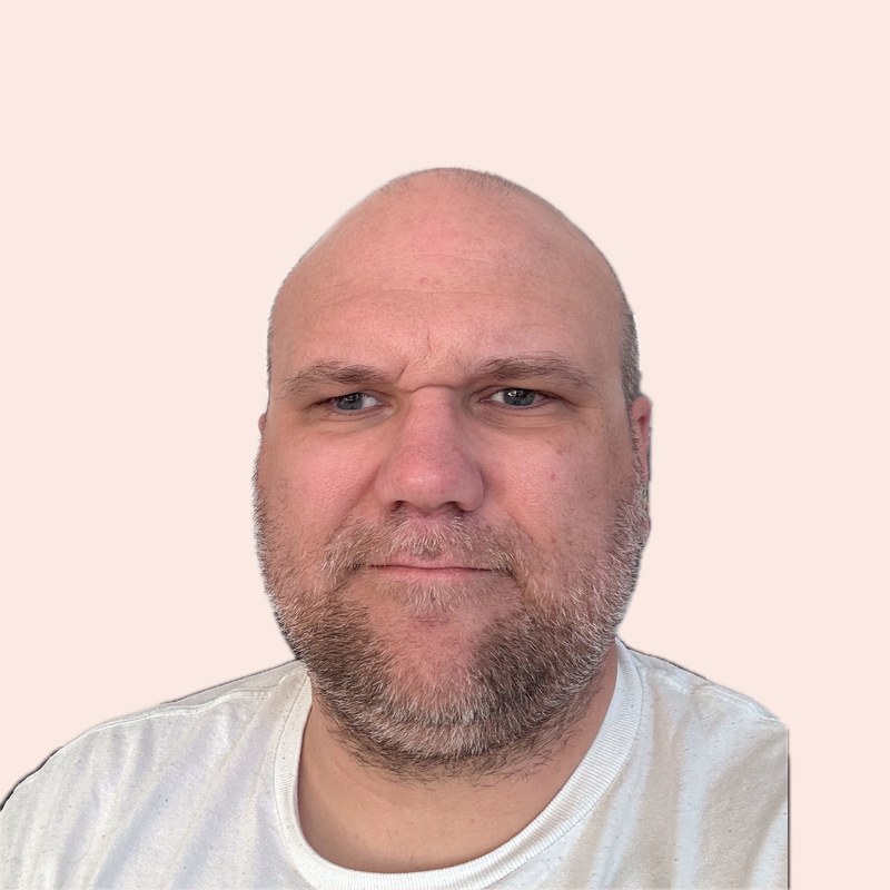 Alexander Ramm blickt mit leichtem Lächeln in die Kamera. Er hat einen kurzen grauen Bart und trägt ein helles T-Shirt.